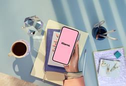 Hand holding phone showing Klarna logo over a desk with coffee and notebooks